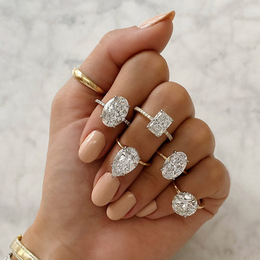 Hand showcasing five diamond engagement rings in different shapes, including oval, cushion, round, and radiant cuts, set on gold bands.